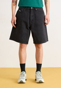 Black denim shorts with a loose fit, featuring front pockets, stitched hem, and a logo patch on the waistband. Paired with gray sneakers and black socks.