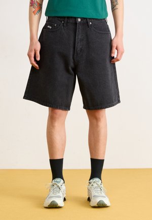 Shorts en denim noir avec une coupe décontractée, dotés de poches avant, d'un ourlet cousu et d'un patch logo sur la ceinture. Associés à des baskets grises et des chaussettes noires.