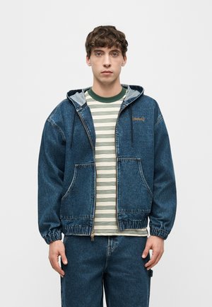 Giovane uomo in piedi rivolto verso avanti che indossa una giacca con cappuccio in denim blu sopra una camicia bianca a righe verdi e pantaloni coordinati in denim blu.