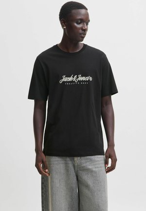 Mand iført sort Jack & Jones t-shirt og lysegrå jeans, står og kigger ned mod sin venstre side mod en ensfarvet baggrund.