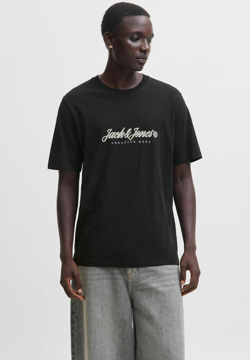 Άνδρας με μαύρο t-shirt Jack & Jones και ανοιχτό γκρι τζιν, στέκεται και κοιτάζει προς τα κάτω αριστερά σε απλό φόντο.