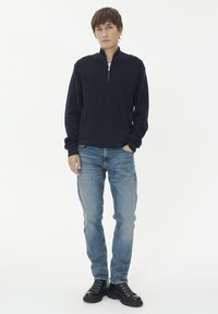 Mornarska zip-svetilka, sproščen kroj, rebrasti manšeti. Ujema se s svetlo modrimi kavbojkami in črnimi vezalnimi čevlji, stoji na beli podlagi.