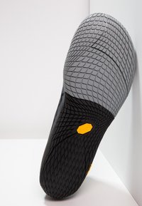 Suola della scarpa nera e grigia con una superficie testurizzata e decorata. Presenta un logo Vibram giallo e bordi del tallone arrotondati.