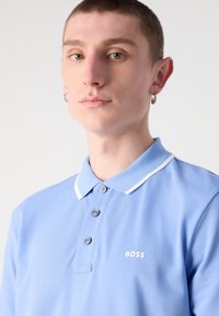 Svijetloplava polo majica s pamučnom teksturom, tri gumba i bijelim obrubom ovratnika. Ima mali bijeli 'BOSS' logo na prsima.