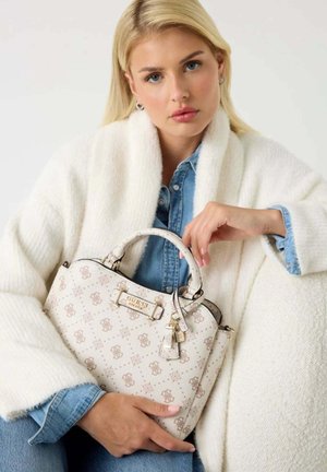 Femme blonde portant un manteau blanc texturé et une chemise en denim, tenant un sac à main beige à motifs Guess avec des accents dorés.