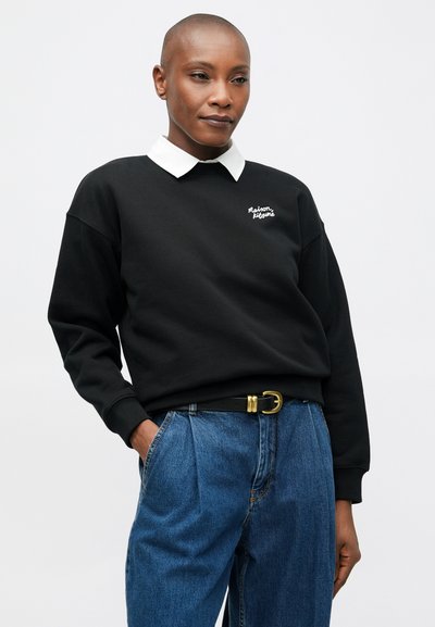 Persona que lleva un sudadera negra con cuello blanco, jeans de cintura alta azules y un cinturón negro con hebilla de oro, de pie con las manos en los bolsillos.