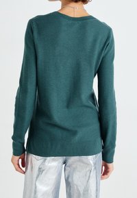 Personne portant un pull à manches longues texturé couleur sarcelle et un pantalon argenté brillant, vue de dos sur un fond clair uni.