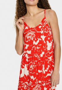 Sarkans un krēmkrāsas rakstains kleita ar V-veida izgriezumu un plānām siksnām, ar brīvu piegriezumu un savilktiem elementiem tuvāk viduklim.