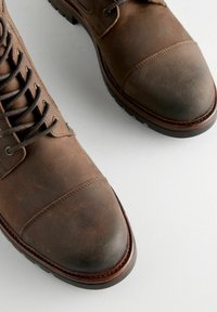 Botas de tobillo de cuero marrón con punta redondeada, diseño tipo cordones, acentos cosidos y suela de goma. Acabado texturizado con un ligero desgaste visible.
