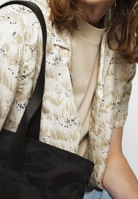 Camicia a maniche corte con motivo floreale beige e accenti blu, sovrapposta a una t-shirt color sabbia chiara; borsa a tracolla nera con la tracolla visibile.