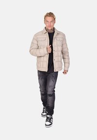 Beige pufferjakke med et quiltet design, båret over en mørk top, kombineret med slidte sorte jeans og sneakers i sort og hvid.