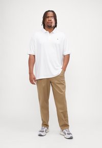 Polo shirt blanc à manches courtes, associé à un pantalon beige et des baskets grises avec des accents noirs et blancs. Design simple et décontracté.