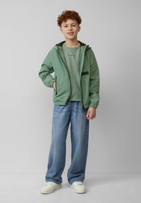 Lichtgroene lichte jack met een capuchon, met ritssluiting aan de voorkant en zijzakken, gecombineerd met een groen t-shirt en losse blauwe jeans. Witte sneakers.