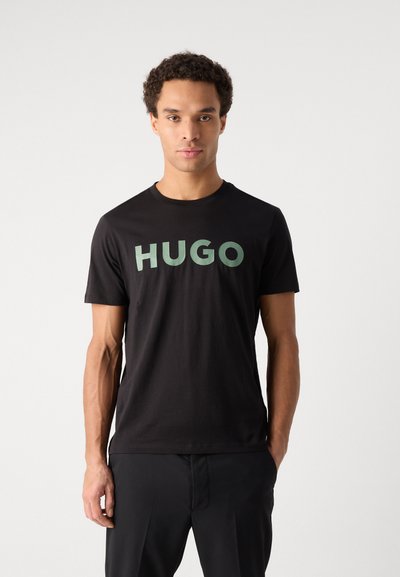 Черна памучна тениска с голям зелен текст "HUGO" отпред. Къси ръкави, кръгло деколте и гладка текстура.