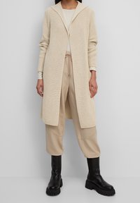 Cardigan lungo beige in maglia con cappuccio a doppio strato, apertura frontale e texture a costine, abbinato a pantaloni tapered beige e stivali neri sopra il ginocchio.