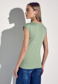 Světlý zelený top s krátkými rukávy a kulatým výstřihem, přiléhavý design a hladká textura, spojený s modrými džínami.
