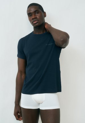 Νέος άνδρας που φοράει μπλε σκούρο μπλουζάκι και λευκά μποξεράκια, στέκεται μπροστά σε απλό λευκό φόντο, με το ένα χέρι στο λαιμό του.