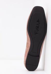 Chaussure plate en suède marron clair avec semelle noire portant le nom de la marque "FURLA" imprimé en dessous.