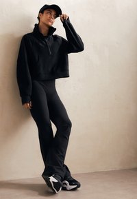 Sweatshirt noir court avec col haut et poche frontale, associé à des leggings noirs moulants et des chaussures de sport noir et blanc.
