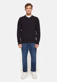 Pulover tricotat bleumarin cu decolteu în V, purtat peste un tricou alb, asortat cu blugi și adidași albi. Prezintă manșete și tiv ribbate.