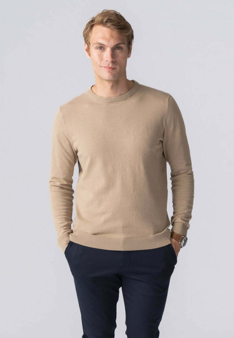 Beige crewneck sweater lavet af blødt materiale, med ribbede manchetter og kant. Parret med mørke bukser, båret afslappet med hænderne i lommerne.