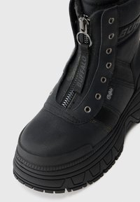 Botín negro de cuero con plataforma y suela gruesa, cremallera frontal etiquetada "BUFFALO" y ojales de metal para los cordones.