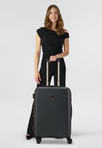 Valise rigide noire avec motif logo embossé, poignée télescopique, quatre roues et accent triangulaire. Texture lisse et forme rectangulaire.