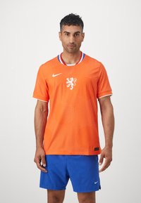 Nike teamshirt in feloranje met een gestippeld patroon, voorzien van een witte leeuwenem en een kraag met blauwe accenten. Gecombineerd met blauwe shorts.