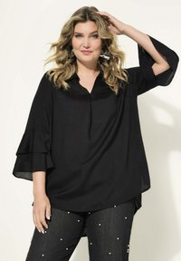 Schwarze Bluse mit ausladenden, geschichteten Ärmeln und V-Ausschnitt. Glattes Material mit lässiger Passform. Kombiniert mit dunklen Jeans, die mit weißen Perlenakzenten verziert sind.