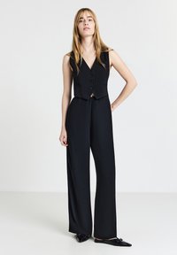 Combinaison noire sans manches avec une taille cintrée, décolleté en V, trois boutons sur le devant et jambes larges, conçue en tissu lisse et texturé.