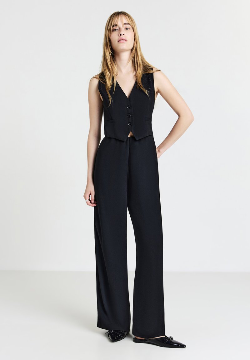 Combinaison noire sans manches avec une taille cintrée, décolleté en V, trois boutons sur le devant et jambes larges, conçue en tissu lisse et texturé.