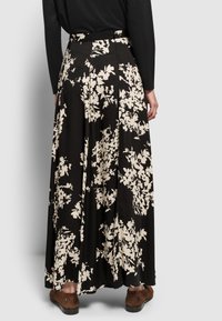 Rückansicht einer Frau, die einen schwarzen Maxirock mit weißen Blumenmuster, ein schwarzes Langarmoberteil und braune Schuhe mit Leopardenmuster trägt.