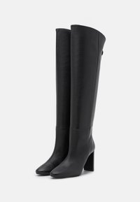 Bottes noires en cuir montantes avec une finition lisse et élégante, bout carré et talon bloc épais. Présente des coutures sur le bord supérieur.