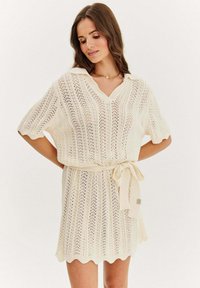 Dzianinowa sukienka z kremowej włóczki, z dekoltem w kształcie V, krótkimi rękawami i wiązanym paskiem w talii. Posiada falbaniaste krawędzie oraz teksturowany wzór.