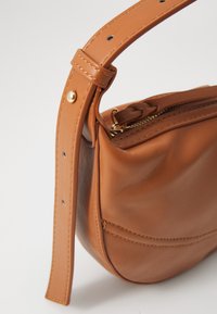 Bolso de cuero marrón claro con forma curva, que presenta un bolsillo con cremallera, una textura suave y una correa larga y ajustable con accesorios dorados.