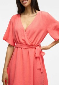 Korálově růžové zavinovací šaty s krátkými rukávy, výstřihem do V, texturovanou tkaninou a sladěným pásem v pase. Charakterizované nařasenými záhyby.
