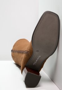 Botte en daim marron avec un bout arrondi, une fermeture éclair latérale et une semelle plate grise. Présente un accent en bois brun foncé sur le talon. Taille 37.