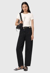 Jean en denim noir taille haute, coupe ample, associé à un T-shirt ajusté crème avec du texte, et un petit sac bandoulière noir. Chaussures noires texturées.