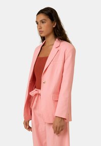 Hellrosa Leinenblazer mit einer Knopfverschluss, Reverskragen und zwei Seitentaschen. Kombiniert mit einem rostfarbenen, gerippten Oberteil.