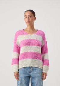 Maglione a righe rosa e bianche, con scollatura a V, vestibilità ampia, maniche lunghe e orlo a costine. Indossato con jeans azzurri a vita alta.