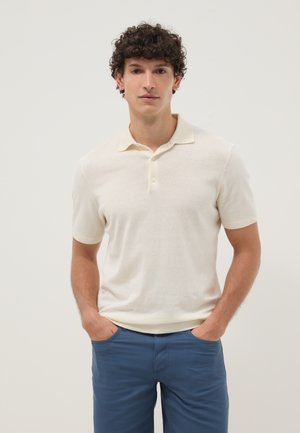 Hombre joven con cabello rizado y oscuro, vistiendo una camiseta tipo polo de manga corta color crema y pantalones azules, con las manos en los bolsillos, sobre un fondo claro y liso.