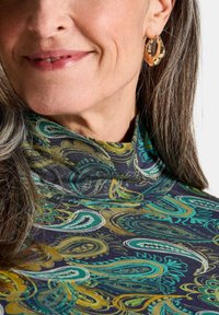 Frau mit langen grauen Haaren, die goldene Creolen trägt und ein buntes Turtleneck-Oberteil mit Paisley-Muster anhat, lächelt dezent.
