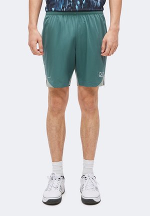 Homme portant un short de sport vert, des chaussettes de sport blanches et des chaussures de course blanches, debout avec les bras détendus le long du corps.