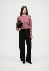 Svetlo roza rebrast cropped top z okroglim izrezom in gumbnim zapiranjem, v kombinaciji s črnimi širokimi hlačami. Model drži črno torbico.