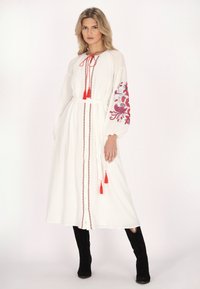 Weißes langes Kleid mit roter Stickerei an den Ärmeln, gebundenem Taillenbereich und Quasten. Verfügt über eine geknöpfte Vorderseite und lange, voluminöse Ärmel. Getragen mit schwarzen Stiefeln.