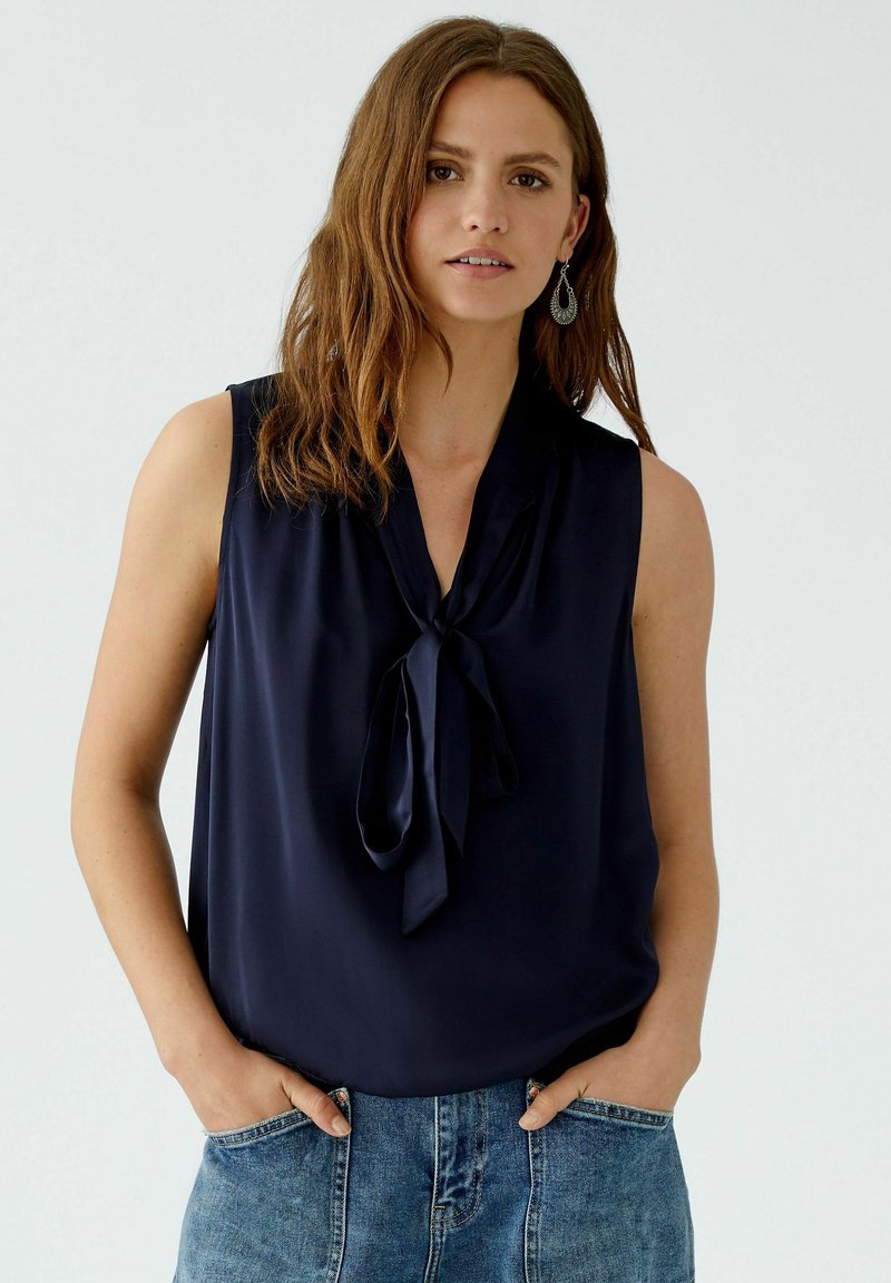 Navyfarbige ärmellose Bluse aus glattem Stoff mit einem Verschluss am Hals. Kombiniert mit Jeansshorts, die eine lockere Passform und ein minimales Design zeigen.