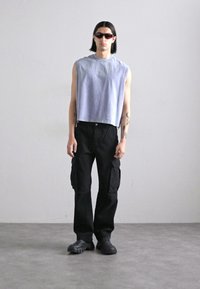 Grå ermeløs crop-top, svarte cargo-bukser med lommer, og svarte sneakers. Modell som har på seg mørke solbriller, står på en nøytral bakgrunn.