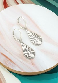 Silver droppformade örhängen med lager av utskurna mönster. Slät yta med en glänsande finish, visad på en marmorerad rund bakgrund.