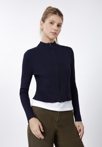 Marineblå ribbestrikket genser med glidelås, høy hals og lange ermer, kombinert med en hvit topp og olivengrønne bukser. Jevn tekstur.