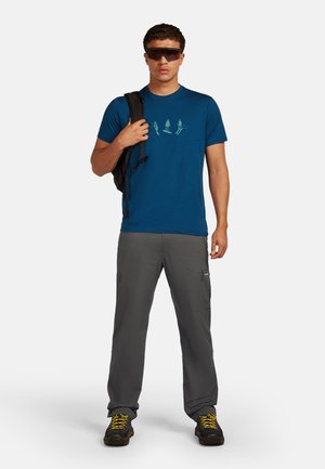 Man in een blauwe graphic t-shirt, grijze broek, wandelschoenen met gele veters, zonnebril en met een zwarte rugzak over één schouder.